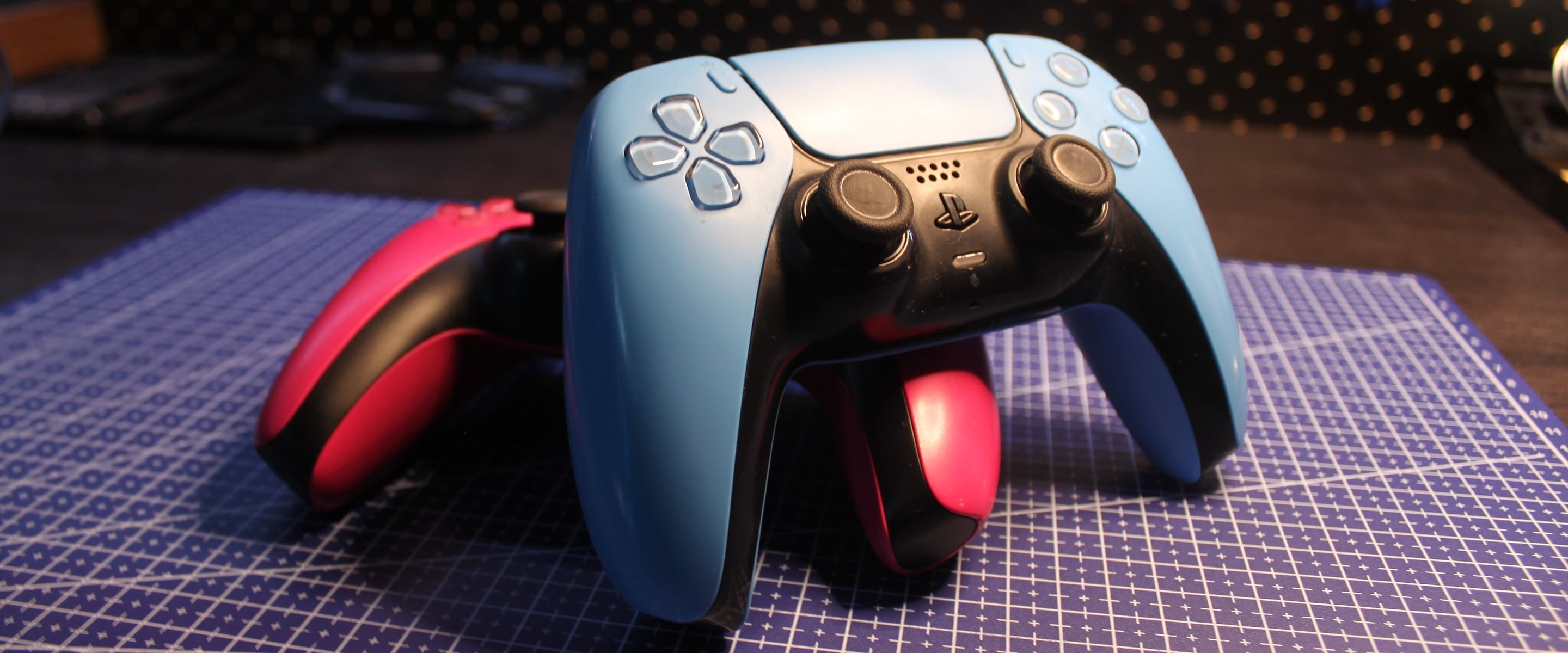 A blue DualSense controller in front of two pink ones on the workbench, with the thumbsticks and triggers we upgrade marked